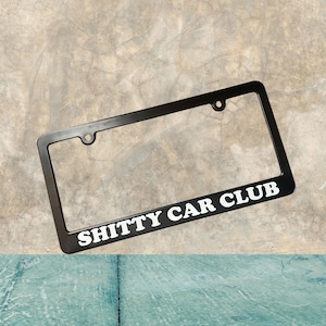 Puede incluir: Marco de matrícula de metal negro con texto blanco que dice "Shitty Car Club".