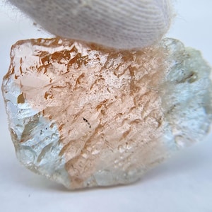 May include: A rough gemstone with a translucent appearance. The stone has a mix of peach, light brown, and pale blue hues. The surface has a textured, crystalline appearance. The gemstone is held by a white fabric.