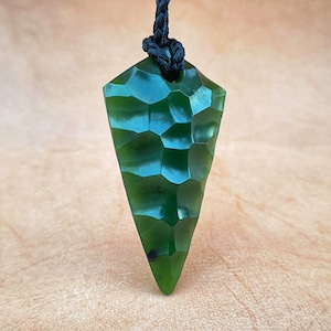 May include: A dark green jade pendant in the shape of a spear point, with a textured surface. The pendant is hanging from a black cord.