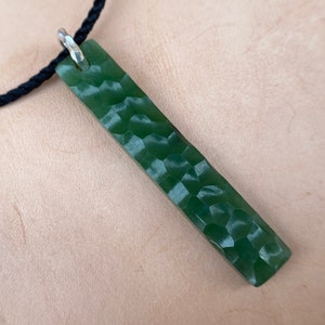 May include: A green jade pendant with a textured surface hangs from a black cord necklace.