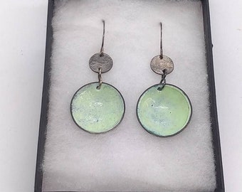 May include: A pair of earrings with a light green enamel finish and a textured silver-tone metal. The earrings are in a black box with the text "SARANNE CONTEMPORARY JEWELLERY". The earrings have a circular design.