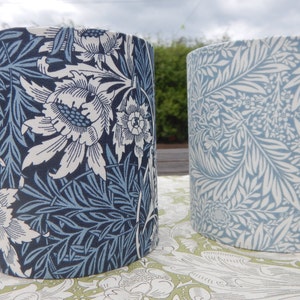 May include: Two cylindrical lampshades with floral patterns. The lampshade on the left has a blue and white floral pattern with large white flowers. The lampshade on the right has a blue and white floral pattern with smaller white flowers.