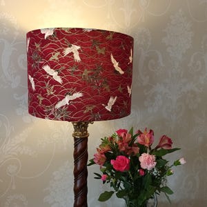 May include: A table lamp with a burgundy lampshade decorated with white cranes. The lamp has a dark brown, twisted wooden base and a gold-coloured top. A vase of pink and red flowers is next to the lamp.