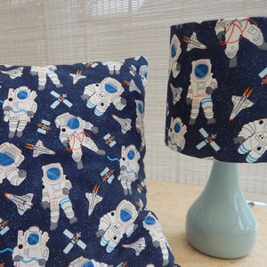 Op de afbeelding: Een blauwe en witte lampenkap en kussen met astronauten, ruimteschepen en sterren. De lampenkap staat op een lichtblauwe lampenvoet.