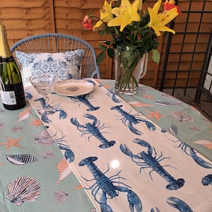 May include: A white table runner with a blue lobster and seashell pattern. The runner is laid on a green tablecloth with a similar pattern. A glass of water and a bottle of wine are on the table.