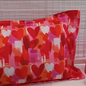 May include: A rectangular pillow and a small cylindrical lampshade, both patterned with red, white, and pink hearts. The pillow is on the left, and the lampshade is next to a small, white candle.