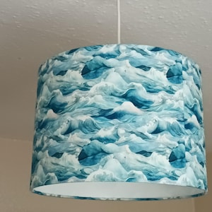 Op de afbeelding: Een cilindrische trommellampenkap met een herhalend patroon van gestileerde oceaangolven in verschillende tinten blauw, groenblauw en wit. De stoffen kap hangt aan een wit snoer en heeft een witte binnenvoering die aan de onderkant zichtbaar is.