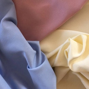 May include: An image showcasing three swatches of fabric in pastel colors. The fabrics are a light blue, a muted rose, and a creamy off-white. The fabric is draped and folded, highlighting its texture and flow.