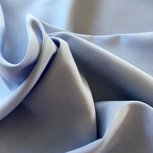 May include: Close-up of light blue fabric with soft folds and subtle shadows. The fabric appears smooth and possibly silky, with a gentle sheen. The overall effect is one of delicate texture and a calming color palette.