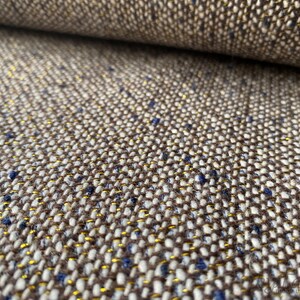 Op de afbeelding: Close-up van een bruine, witte en gouden tweed stof met een subtiel blauwe schijn. De stof heeft een textuur en geweven uiterlijk.