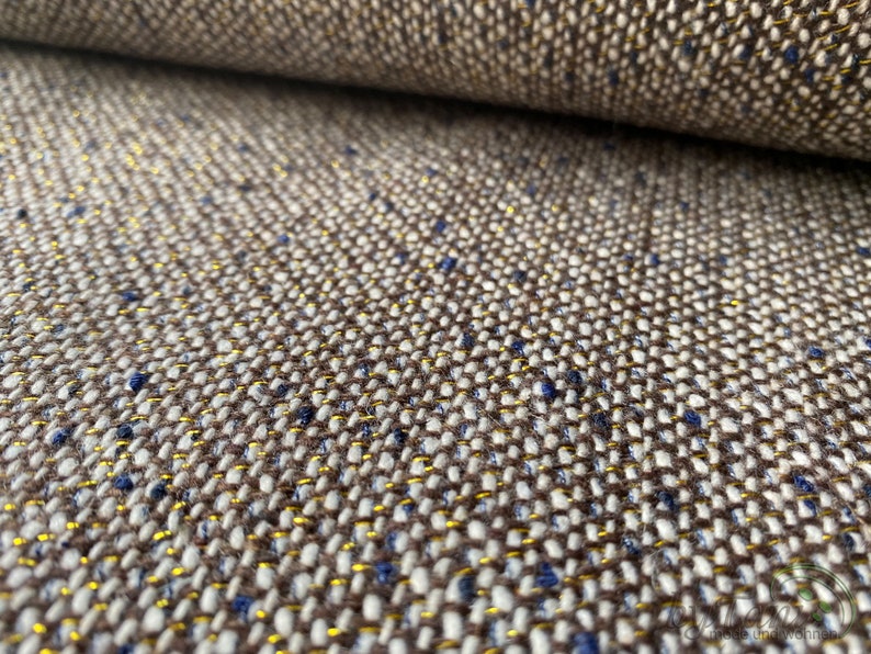 Op de afbeelding: Close-up van een bruine, witte en gouden tweed stof met een subtiel blauwe schijn. De stof heeft een textuur en geweven uiterlijk.