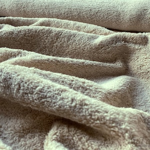 May include: Close-up of a beige, soft, fluffy fabric with a textured surface. The fabric is folded and draped, showing its texture and softness.