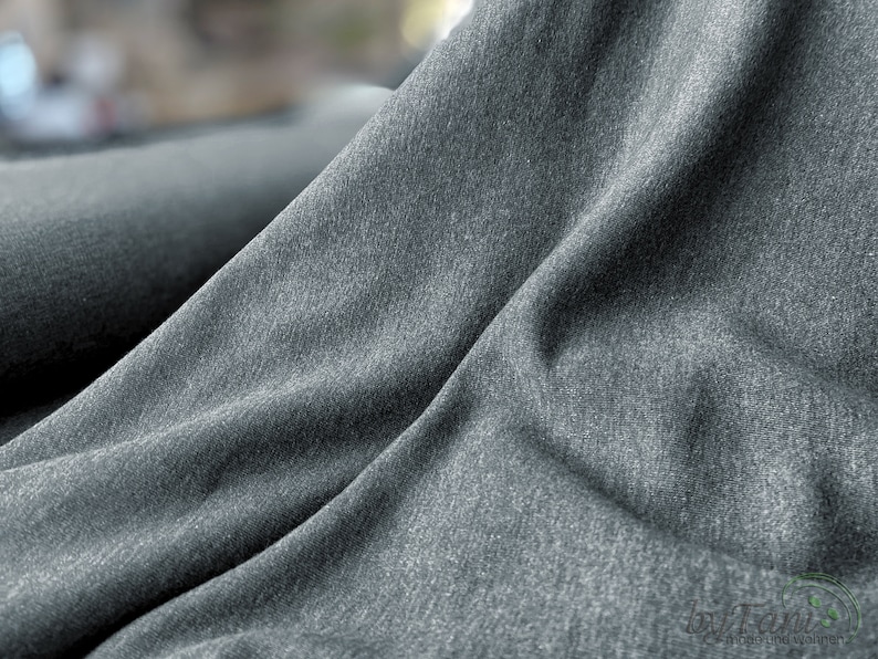 May include: Close-up of a dark gray fabric with a textured surface. The fabric is folded and draped, showing its soft and drapey texture.