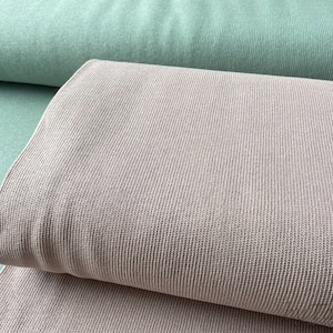 Peut inclure: Gros plan sur deux échantillons de tissu. L'un est vert sauge clair, l'autre est de couleur taupe neutre pliée. Les deux tissus ont une texture côtelée, suggérant un matériau tricoté ou texturé. Les tissus sont probablement pour l'habillement ou la décoration intérieure.