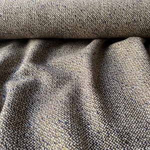 Op de afbeelding: Close-up van een bruine tweed stof met spikkels van gouden en blauwe draad. De stof is geweven in een textuurpatroon en heeft een zacht, luxueus gevoel.