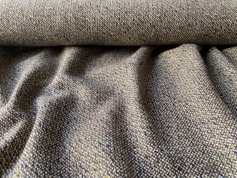 Op de afbeelding: Close-up van een bruine tweed stof met spikkels van gouden en blauwe draad. De stof is geweven in een textuurpatroon en heeft een zacht, luxueus gevoel.