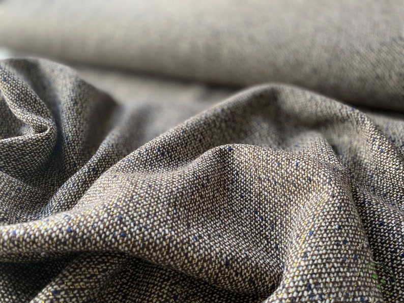 Op de afbeelding: Close-up van een bruine tweed stof met een subtiele glans. De stof heeft een textuur met gouden en blauwe vlekken.