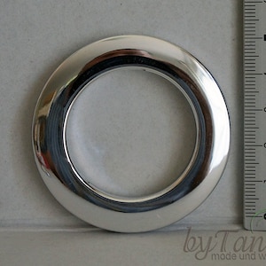 Op de afbeelding: Een glanzende, zilverkleurige gordijnring met een cirkelvormig ontwerp. Een liniaal staat naast de ring en toont metingen in centimeters. De ring is gemaakt van een reflecterend materiaal en de foto is van bovenaf genomen.