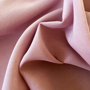 May include: Close-up of a soft, flowing fabric in a muted rose color. The fabric is draped and folded, creating shadows and highlights that emphasize its texture. The image suggests a textile suitable for clothing or home decor.