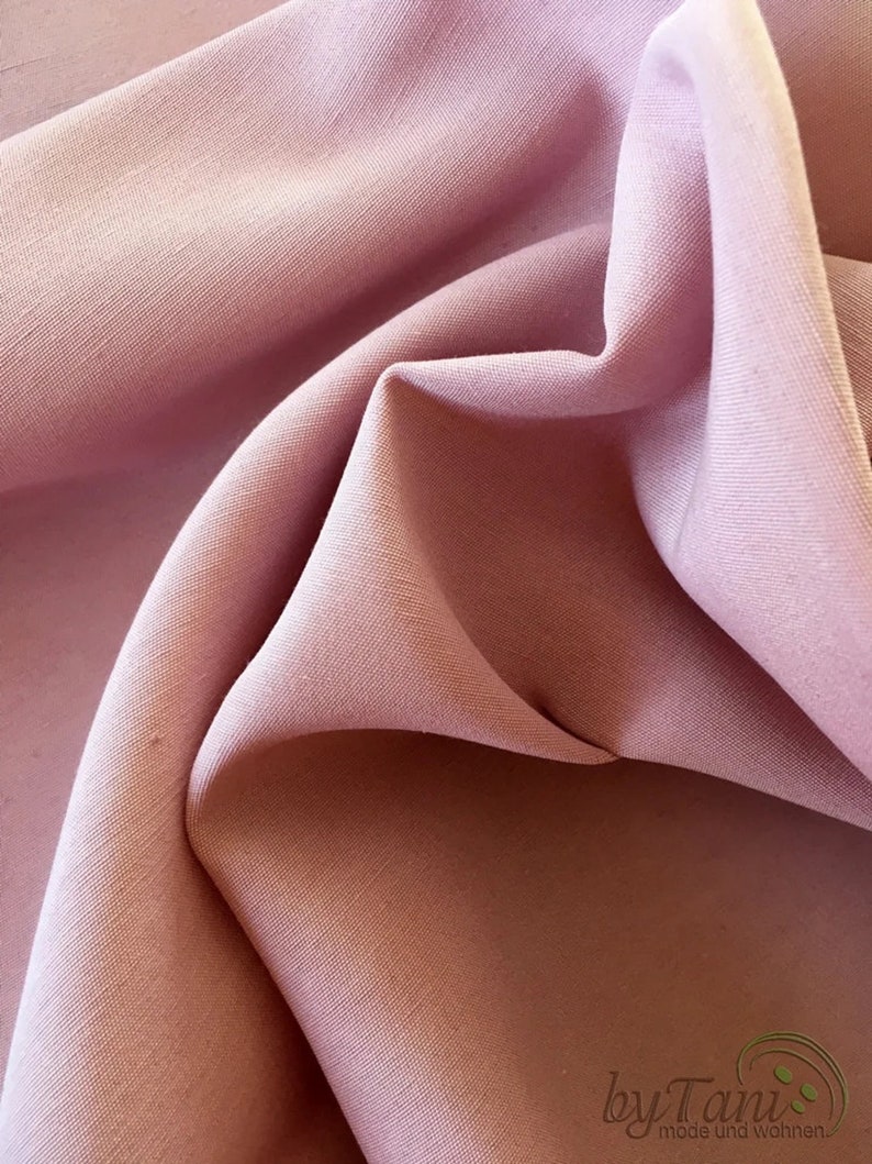 May include: Close-up of a soft, flowing fabric in a muted rose color. The fabric is draped and folded, creating shadows and highlights that emphasize its texture. The image suggests a textile suitable for clothing or home decor.