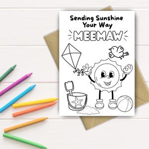 Puede incluir: Una tarjeta blanca con el texto "Sending Sunshine Your Way MEEMAW" e ilustraciones de una cometa, un pájaro, un sol sonriente, un cubo de arena y un balón de playa. Crayones de colores están esparcidos alrededor de la tarjeta.