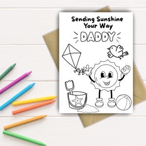 Puede incluir: Una tarjeta en blanco y negro con el texto "Sending Sunshine Your Way DADDY" e ilustraciones de una cometa, un pájaro, un sol sonriente, un cubo de playa, arena y un balón de playa. Los crayones de colores están esparcidos alrededor de la tarjeta.