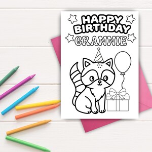 May include: A black and white birthday card with the text "HAPPY BIRTHDAY GRANNIE" and a cartoon raccoon wearing a party hat. The raccoon is holding a balloon and sitting next to a gift box. Colored crayons are scattered around the card.