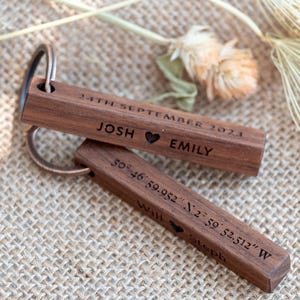 May include: Two wooden keychains with metal rings. The top keychain is engraved with "24TH SEPTEMBER 2023", "JOSH", and "EMILY" with a heart symbol. The bottom keychain has coordinates and the names "Will" and "Steph".