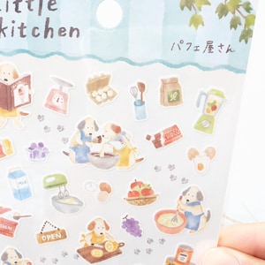 May include: A sticker sheet titled Little Kitchen with Japanese text featuring watercolor illustrations of dogs baking, kitchen appliances, ingredients like milk, eggs, fruit, chocolate, and small paw prints scattered across the clear plastic packaging.