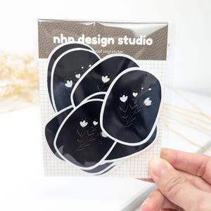Puede incluir: Un paquete de pegatinas de vinilo con un diseño floral. Las pegatinas son negras con ilustraciones florales blancas y un borde blanco. El paquete tiene el texto "nhp design studio" impreso.