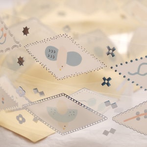 May include: A roll of clear, decorative tape featuring repeating diamond-shaped patterns. Each diamond contains a unique illustration of birds, stars, and other whimsical designs in soft pastel colors. The tape is partially unrolled, revealing the design.