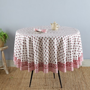 May include: White round tablecloth with a red floral print and a red border. The tablecloth is on a round table with black legs. A small white teapot is on the table.