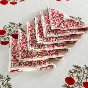 May include: A set of eight white cotton napkins with a red floral print. The napkins are folded in a triangular shape and are arranged in a stack.