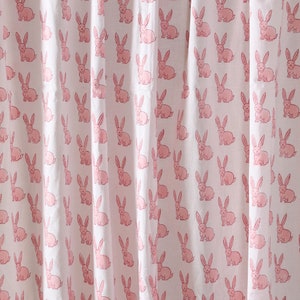 Puede incluir: Tela blanca con un patrón repetitivo de ilustraciones de conejos rosas. Los conejos están representados en un estilo de dibujos animados simple, creando un diseño lúdico. La tela probablemente se utiliza para cortinas u otros artículos de decoración del hogar.