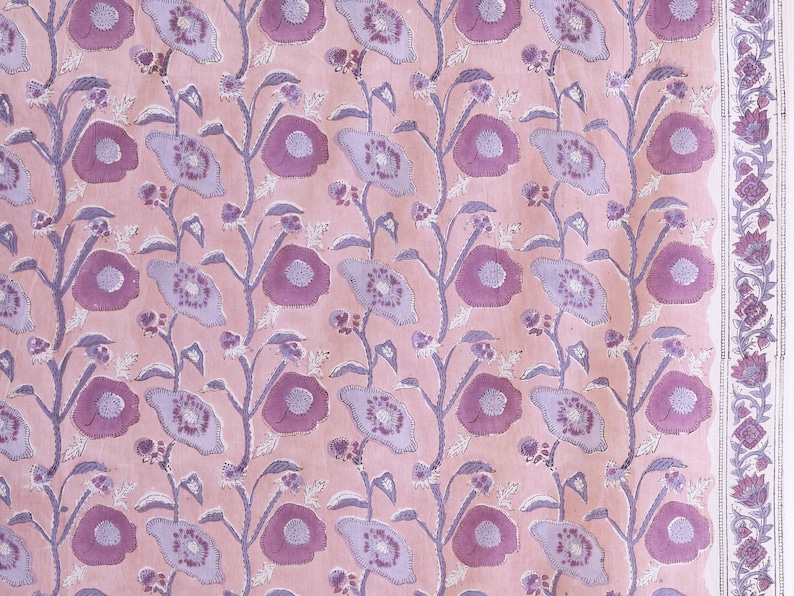 May include: A pink fabric with a repeating pattern of purple flowers and leaves. The fabric has a white border with a floral design.