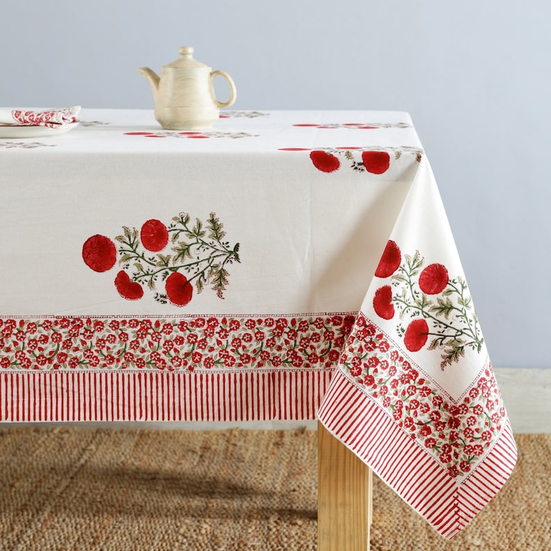May include: White tablecloth with a red floral print. The tablecloth has a red and white striped border. A small white teapot sits on the tablecloth.