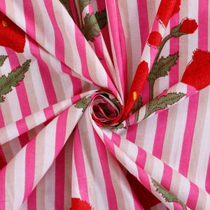 Op de afbeelding: Roze en witte gestreepte stof met een bloemenprint van rode klaprozen. De stof is gevouwen en gedraaid, waardoor het patroon en de textuur zichtbaar zijn.