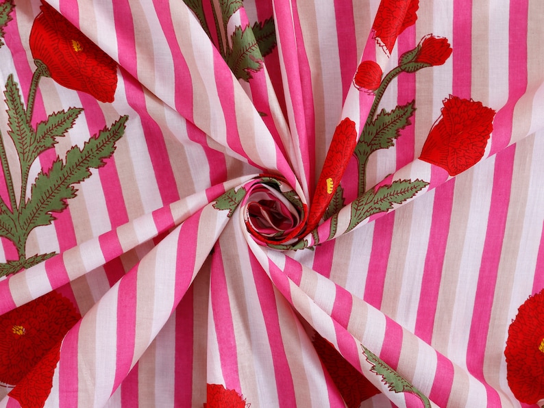 Op de afbeelding: Roze en witte gestreepte stof met een bloemenprint van rode klaprozen. De stof is gevouwen en gedraaid, waardoor het patroon en de textuur zichtbaar zijn.