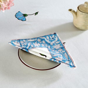 Pode incluir: Um guardanapo de algod&atilde;o azul e branco com um padr&atilde;o floral, dobrado e colocado em um prato branco com uma borda marrom.