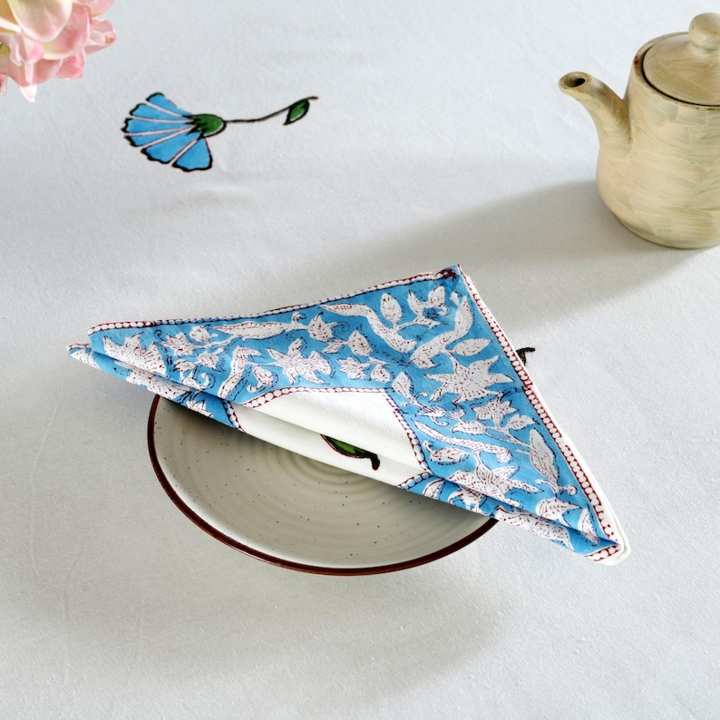 Pode incluir: Um guardanapo de algod&atilde;o azul e branco com um padr&atilde;o floral, dobrado e colocado em um prato branco com uma borda marrom.