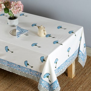 Pode incluir: Toalha de mesa de algod&atilde;o branco com estampa floral azul e borda azul e branca. A toalha de mesa est&aacute; posta sobre uma mesa com uma perna de madeira e um tapete tecido marrom por baixo.