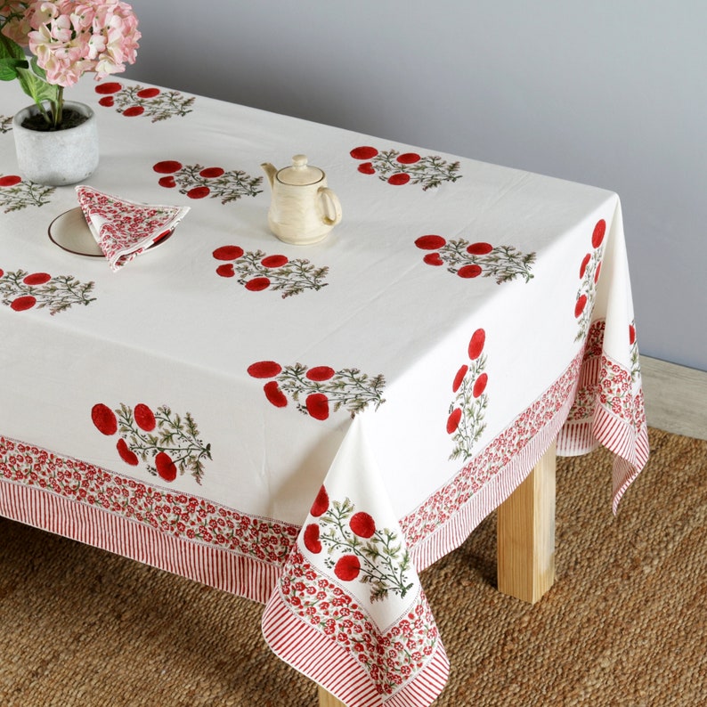 May include: White cotton tablecloth with a red floral print. The tablecloth has a red and white striped border. The tablecloth is laid out on a wooden table with a teapot and a napkin on top.