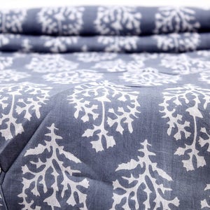 May include: Close-up of a folded fabric with a repeating white tree-like pattern on a blue background. The fabric is textured and appears to be cotton or linen. The pattern is evenly spaced across the material.