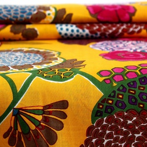 May include: Yellow fabric with a colorful floral pattern. The pattern features large flowers with pink, purple, and green accents, as well as smaller geometric shapes in brown, red, and yellow.