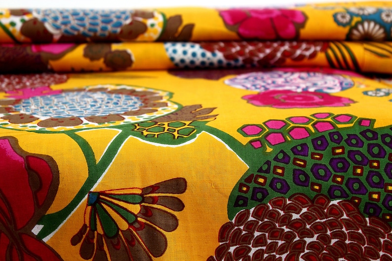 May include: Yellow fabric with a colorful floral pattern. The pattern features large flowers with pink, purple, and green accents, as well as smaller geometric shapes in brown, red, and yellow.