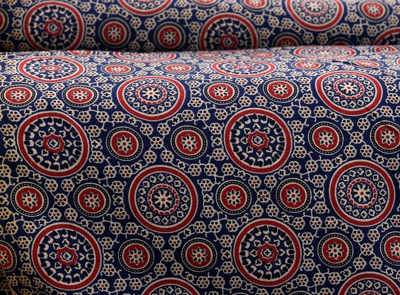 Op de afbeelding: Een blauwe stof met een herhalend patroon van rode, witte en zwarte cirkels. De cirkels hebben ingewikkelde ontwerpen in hun binnenste.