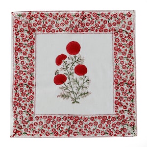 May include: White cotton square table runner with a red floral design. The floral design features a cluster of red flowers with green leaves and stems. The border of the runner is a repeating pattern of small red flowers.