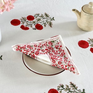 May include: A white cotton napkin with a red floral print. The napkin is folded in a triangle and placed on a white plate. The plate is on a white tablecloth with a red floral print.