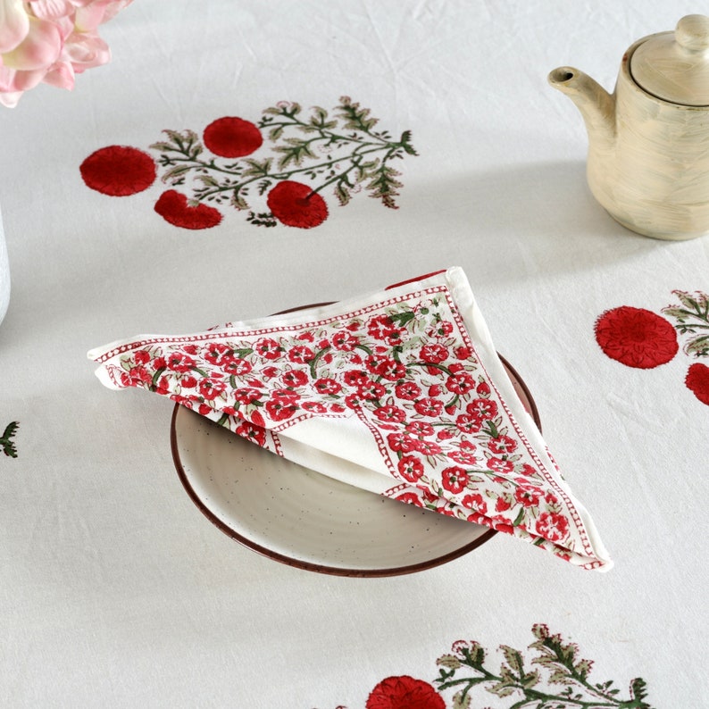 May include: A white cotton napkin with a red floral print. The napkin is folded in a triangle and placed on a white plate. The plate is on a white tablecloth with a red floral print.