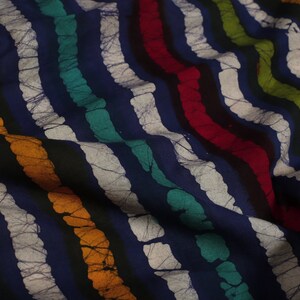 May include: Close-up of a blue fabric with white, black, orange, green, and red stripes. The fabric has a batik pattern with a textured, crinkled look.